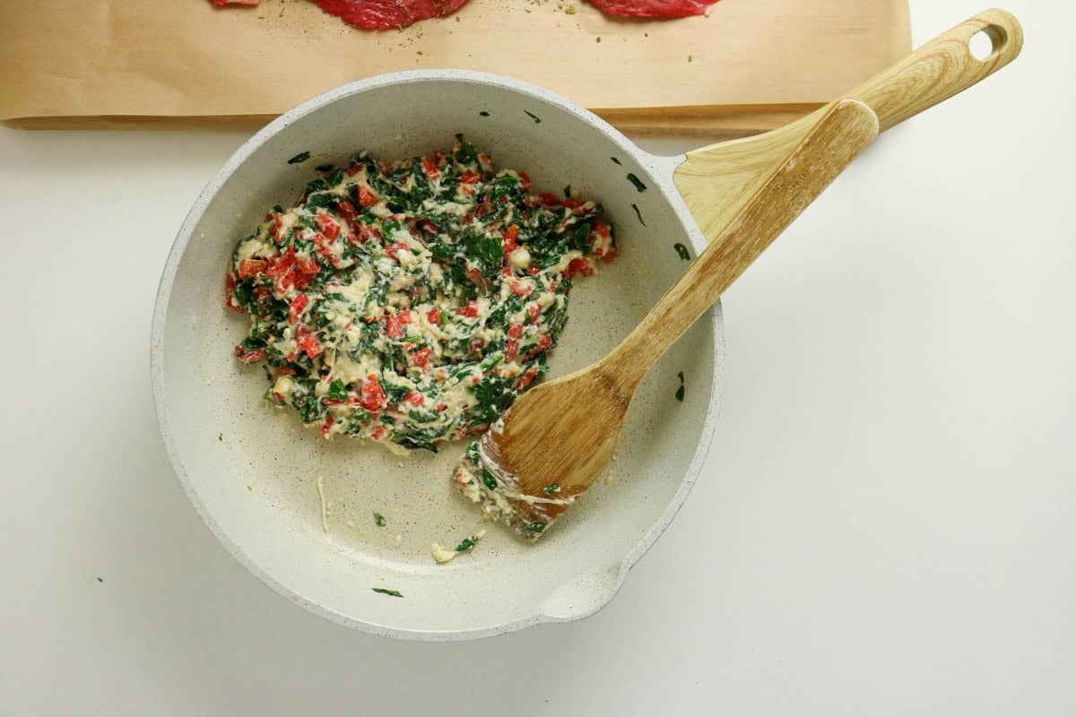 Stirring creamy spinach and red pepper mixture in a pan with a wooden spoon.