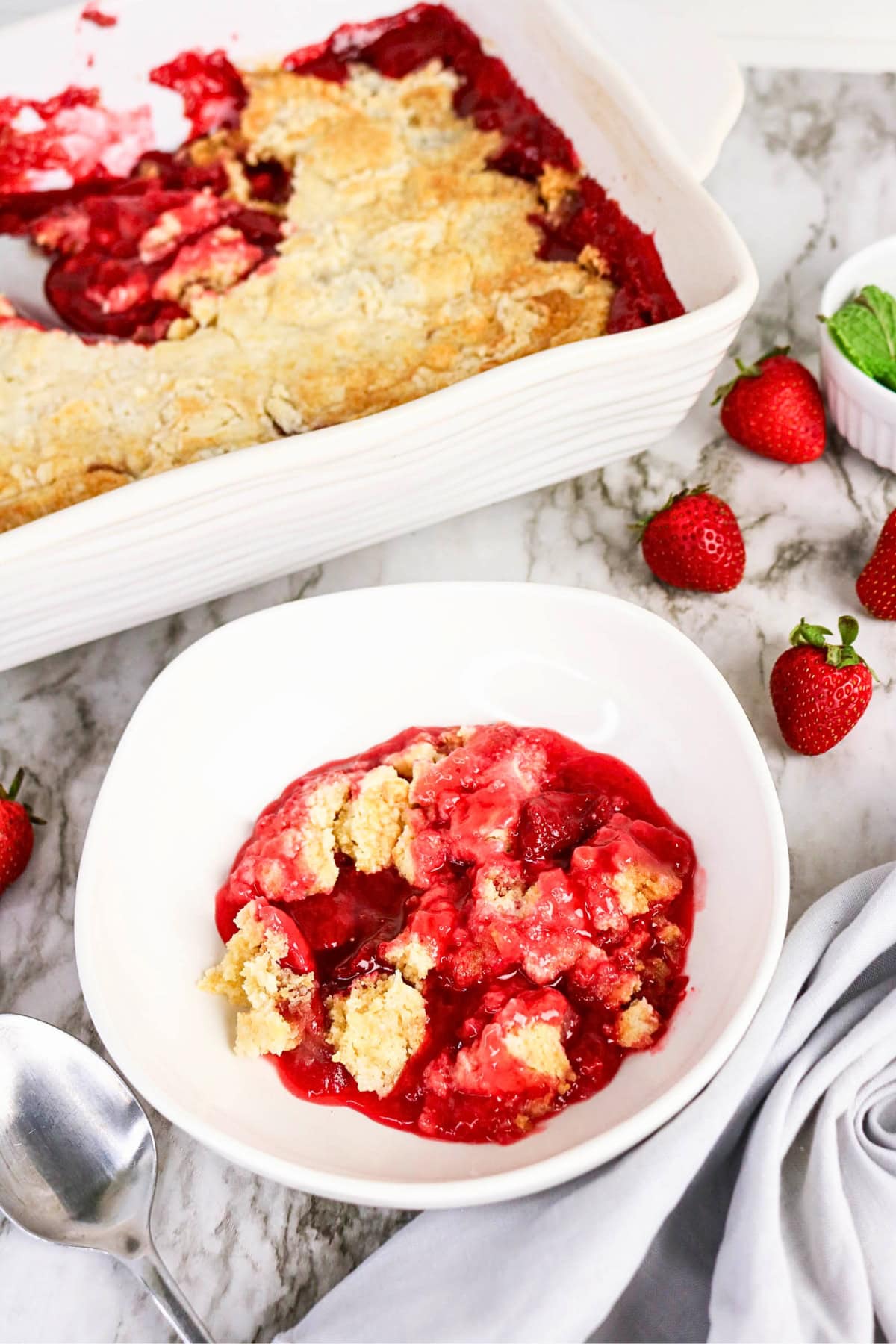 A bowl of strawberry cobbler with fresh strawberries and a baking dish nearby.
