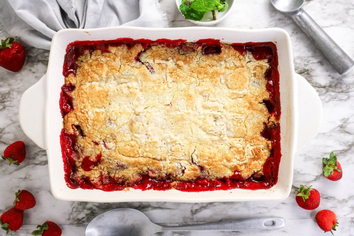 A baked strawberry cobbler in a white dish, surrounded by fresh strawberries.