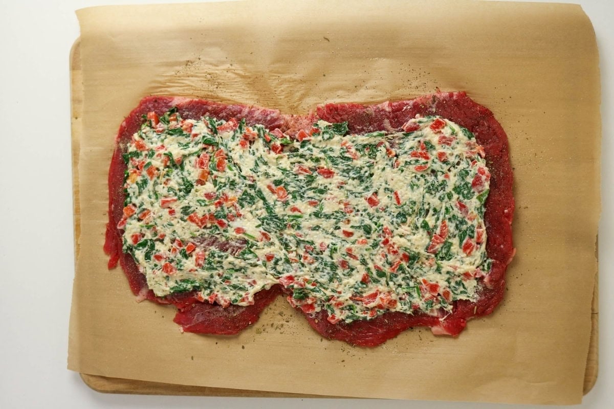 Raw beef topped with a creamy spinach and red pepper mixture on parchment paper.