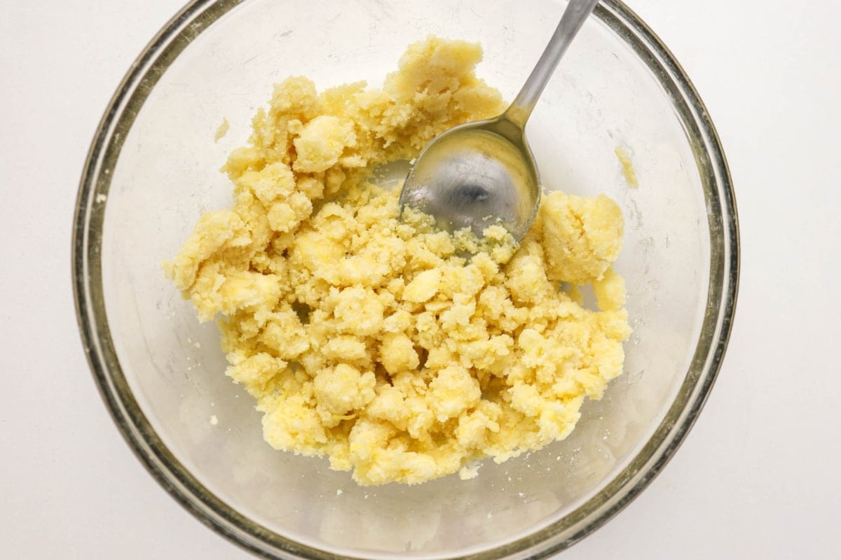 A glass bowl with crumbly dough and a metal spoon inside.
