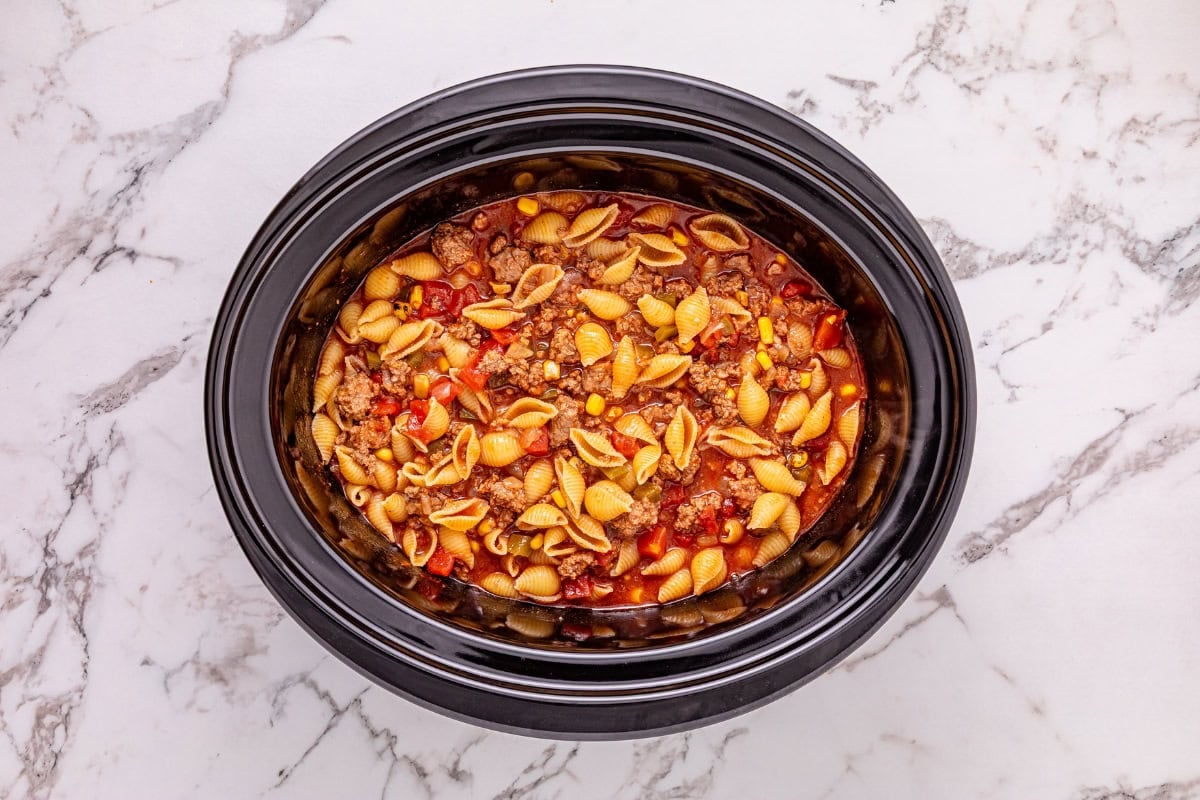 Slow cooker with pasta, ground meat, and tomato sauce on a marble countertop.