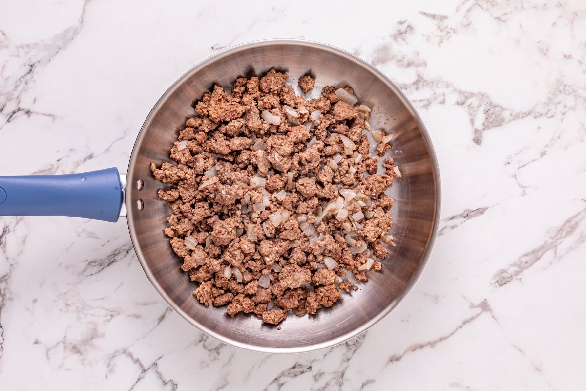 Ground beef and diced onions cooking in a skillet on a marble countertop.