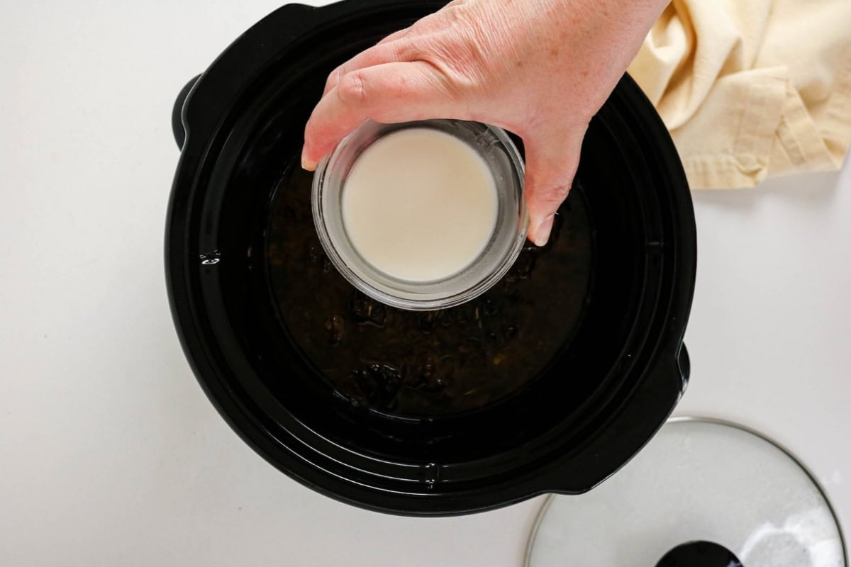 Hand pouring cornstarch from a glass into a black slow cooker with dark liquid inside.
