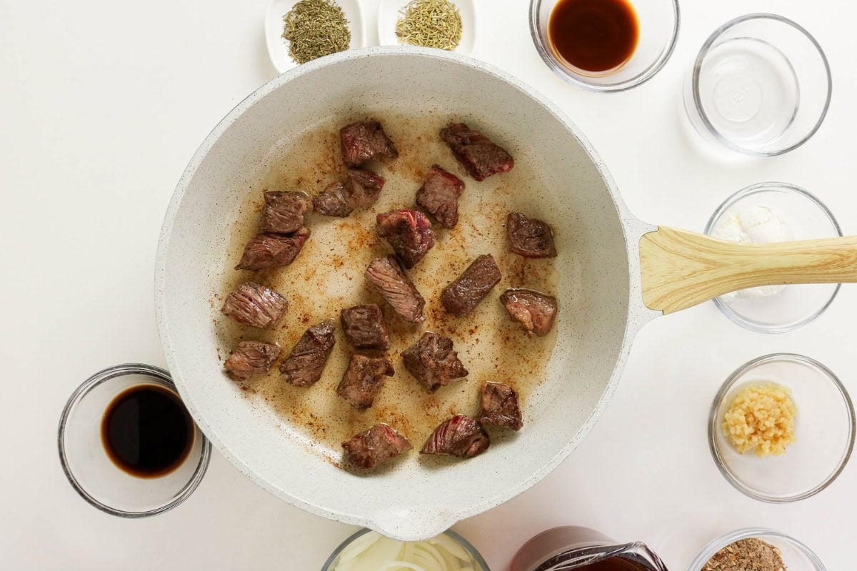 Chunks of beef cooking in a pan, surrounded by seasonings and sauces in bowls.