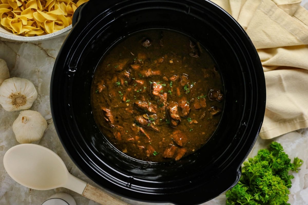 Beef stew in a black slow cooker, surrounded by herbs, garlic, and pasta.