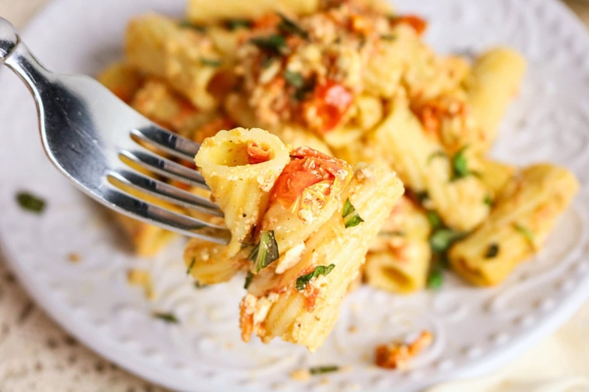 Fork holding rigatoni pasta with tomato, herbs, and cheese over a white plate.