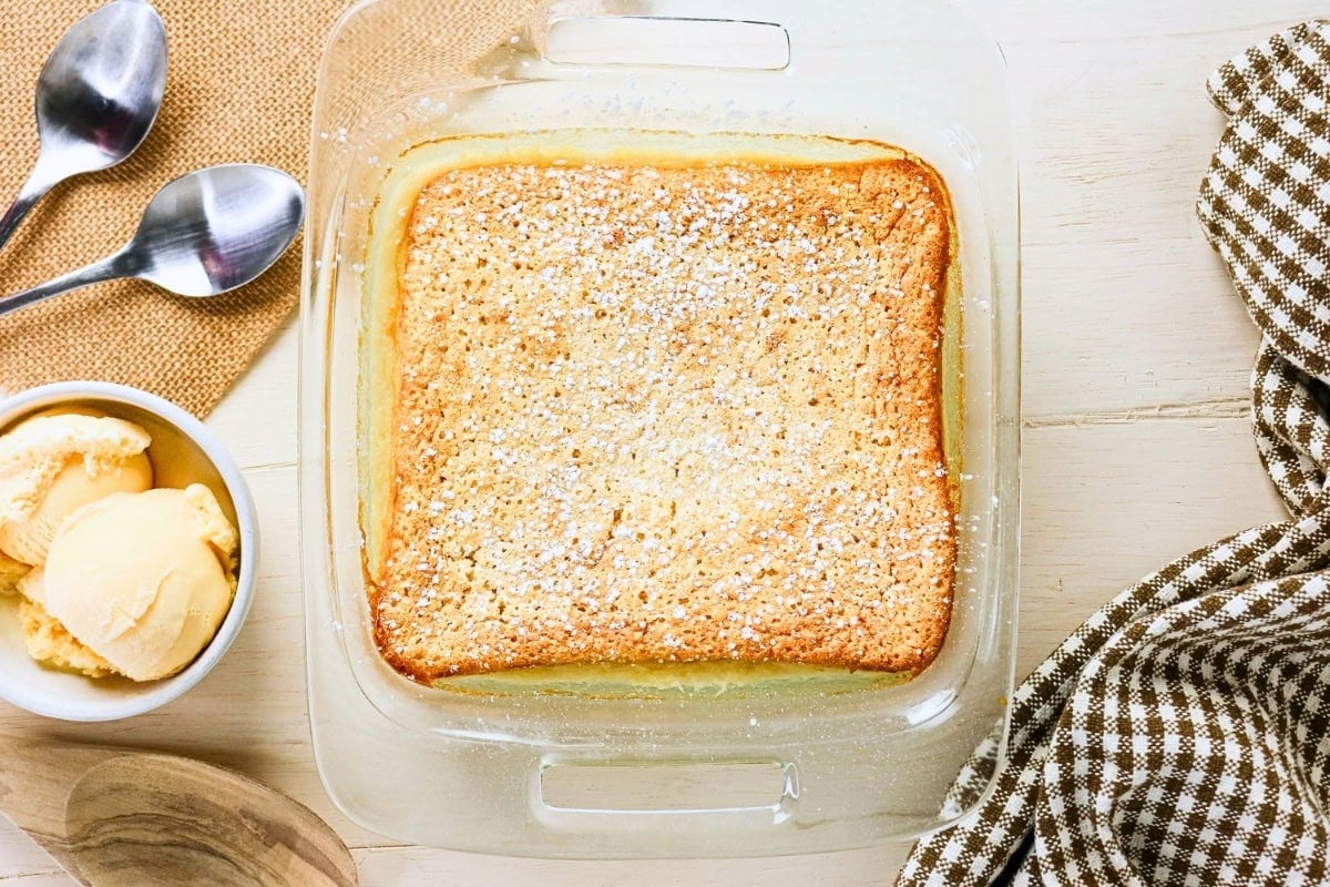 Golden cake in a glass dish, with ice cream and two spoons nearby on a table.