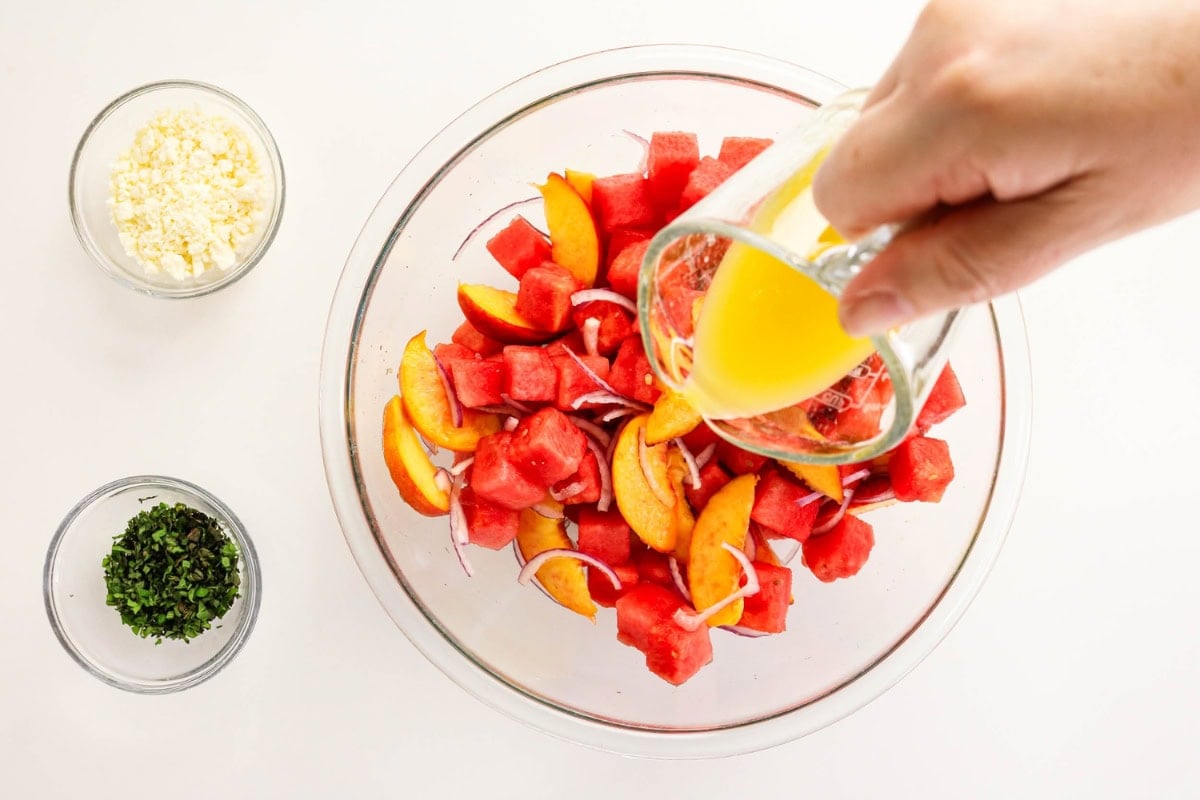 Hand pours orange dressing onto watermelon and peach salad in a glass bowl.