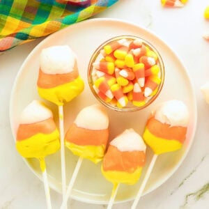 Five candy corn cake pops and a bowl of candy corn on a white plate.