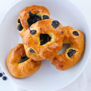 Four blueberry bagels stacked in a white bowl, viewed from above.