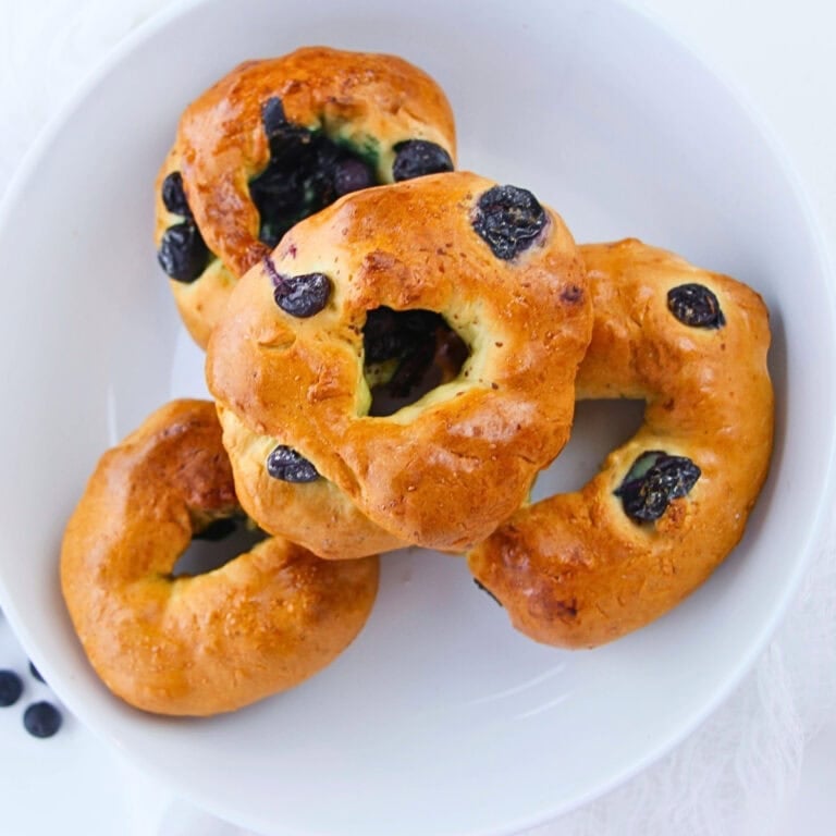Four blueberry bagels stacked in a white bowl, viewed from above.