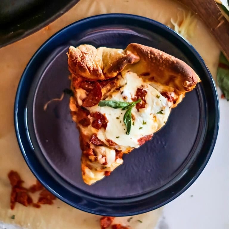 Slice of pizza with cheese and herbs on a dark plate, viewed from above.