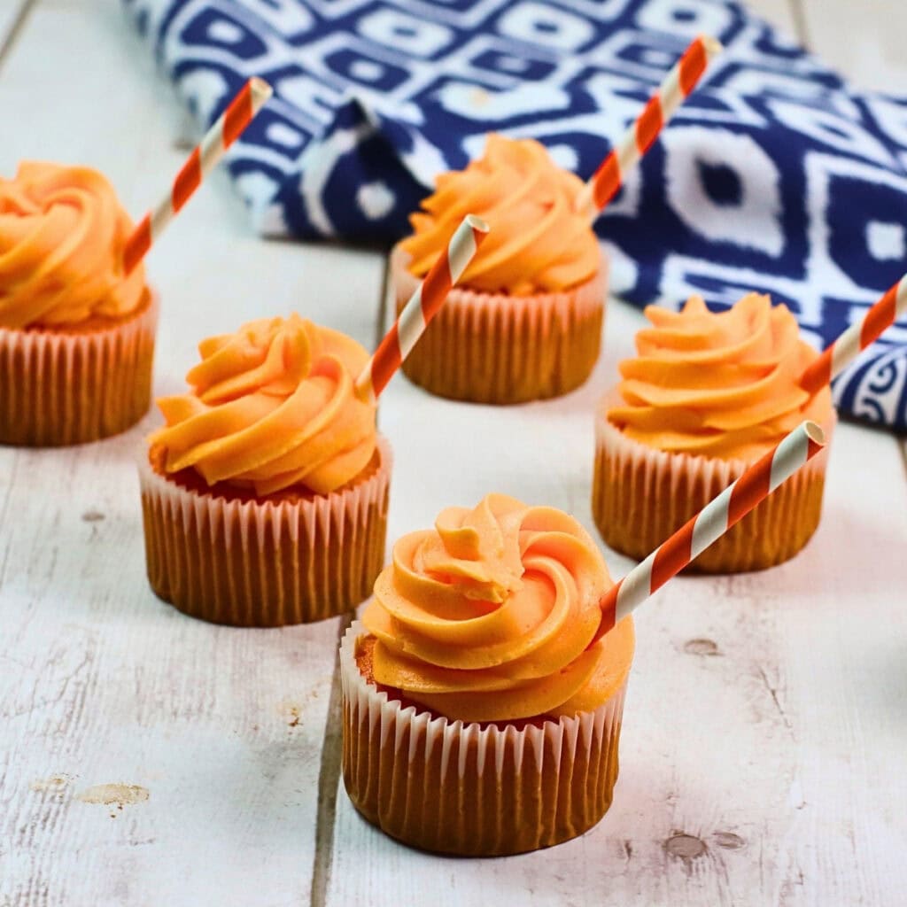 Six cupcakes with orange frosting and striped straws on a wood surface.