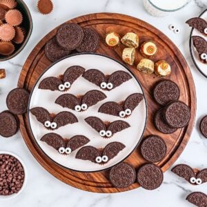 Chocolate cookies decorated as bats with candy eyes on a wooden tray.