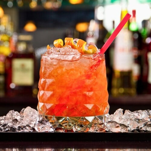 Orange cocktail with ice and a red straw, garnished with orange twists.