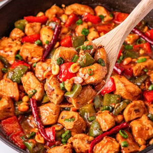 Wooden spoon scooping spicy stir-fried chicken with peppers and peanuts.