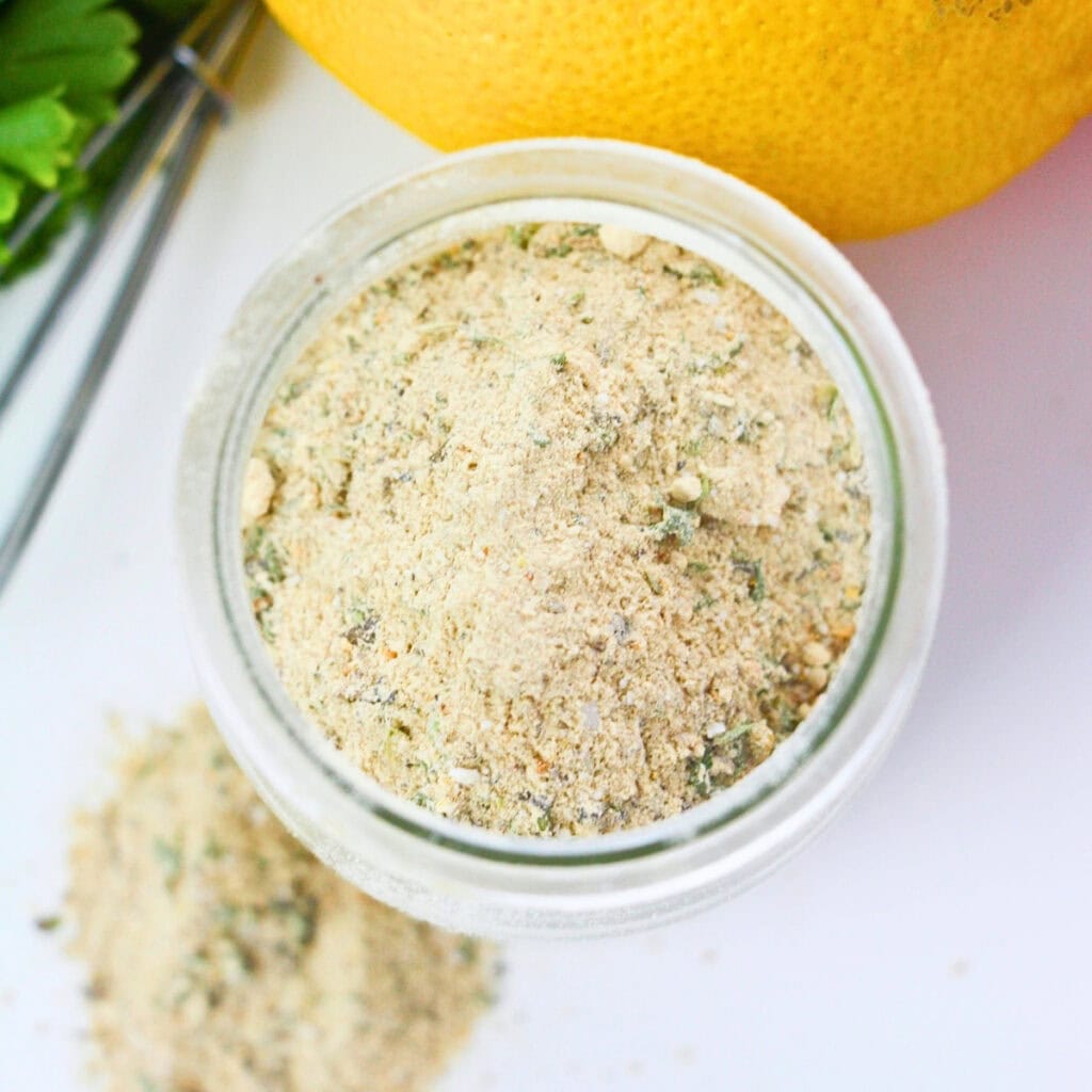 Jar of seasoned breadcrumb mix with spilled crumbs, lemon, and herbs nearby.