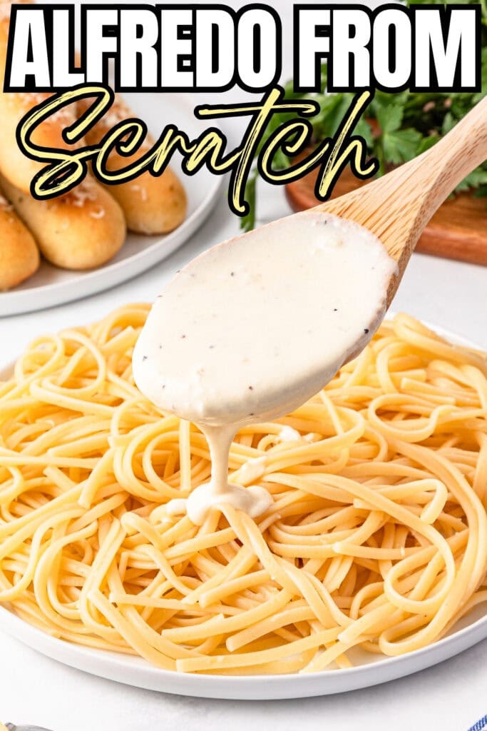 Wooden spoon pours Alfredo sauce over pasta with breadsticks and herbs nearby.