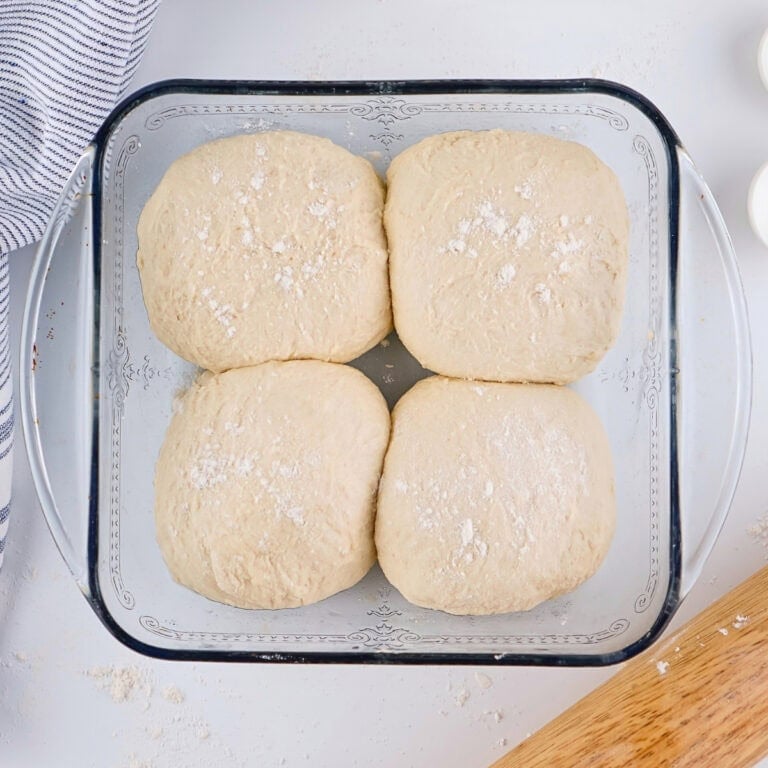 Four dough balls in a square glass dish, sprinkled with flour.
