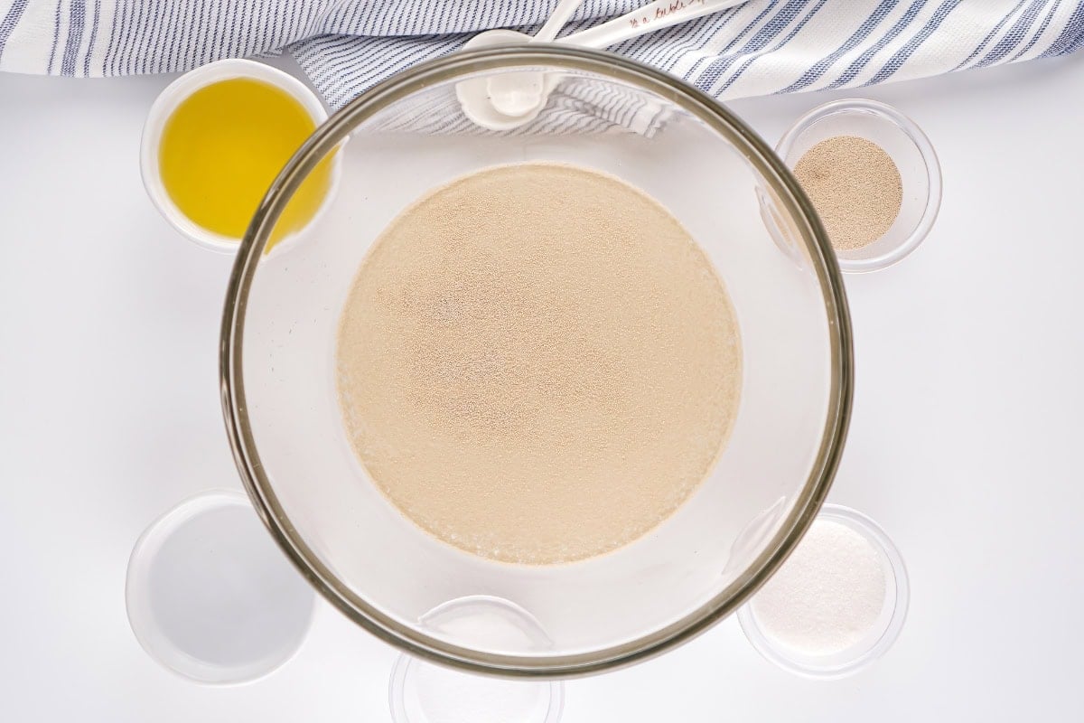 Glass bowl with yeast mixture, surrounded by bowls of ingredients, top view.