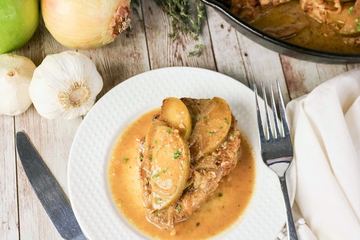 Pork chop with apple slices and sauce on a white plate with fork and knife.