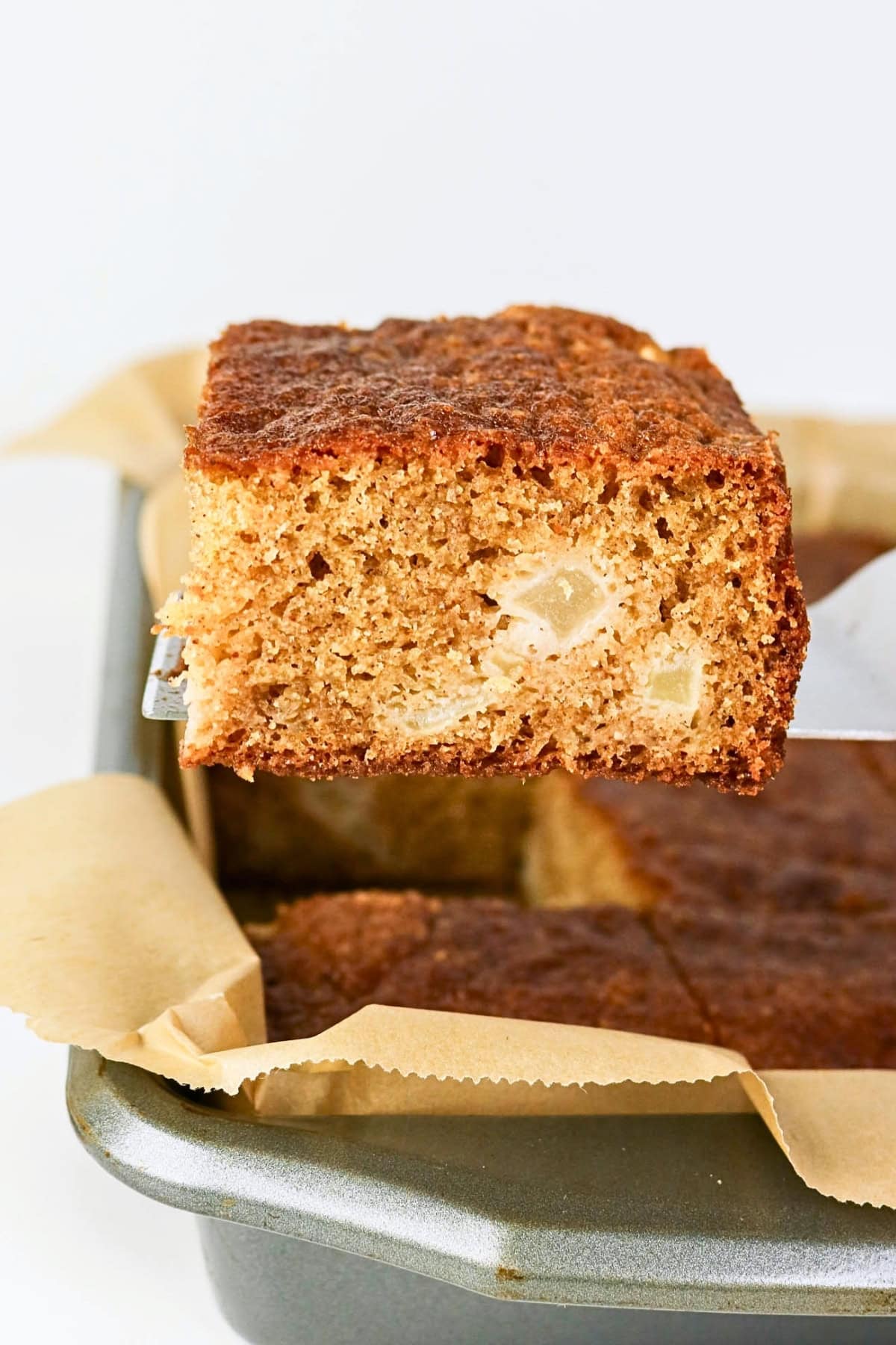 A square piece of brown cake with apple chunks held above a baking pan.