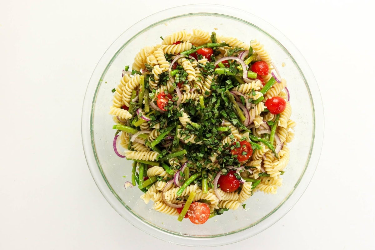 Pasta salad with cherry tomatoes, red onion, herbs, and greens in a glass bowl.