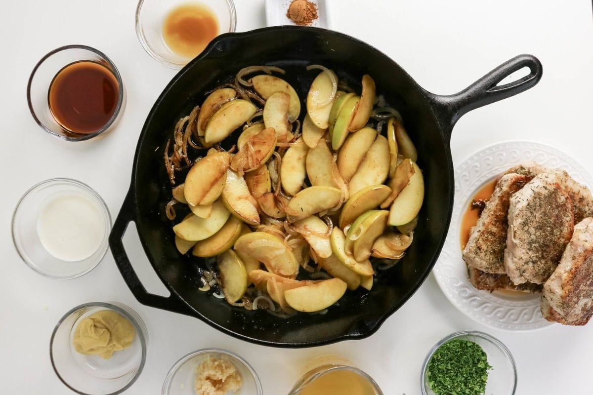 Sliced apples and onions cook in a skillet, surrounded by spices and chicken.