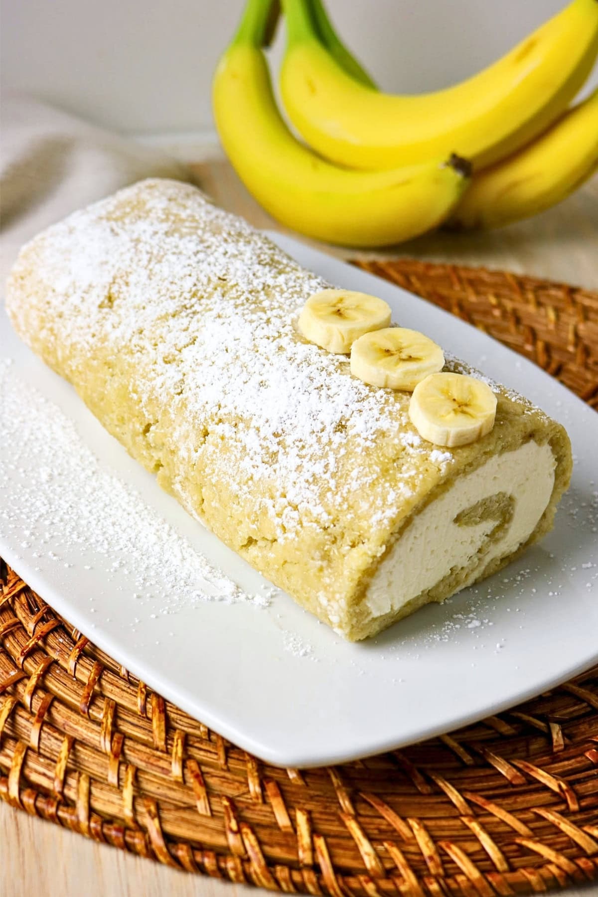 Banana roll cake with powdered sugar, topped with banana slices.