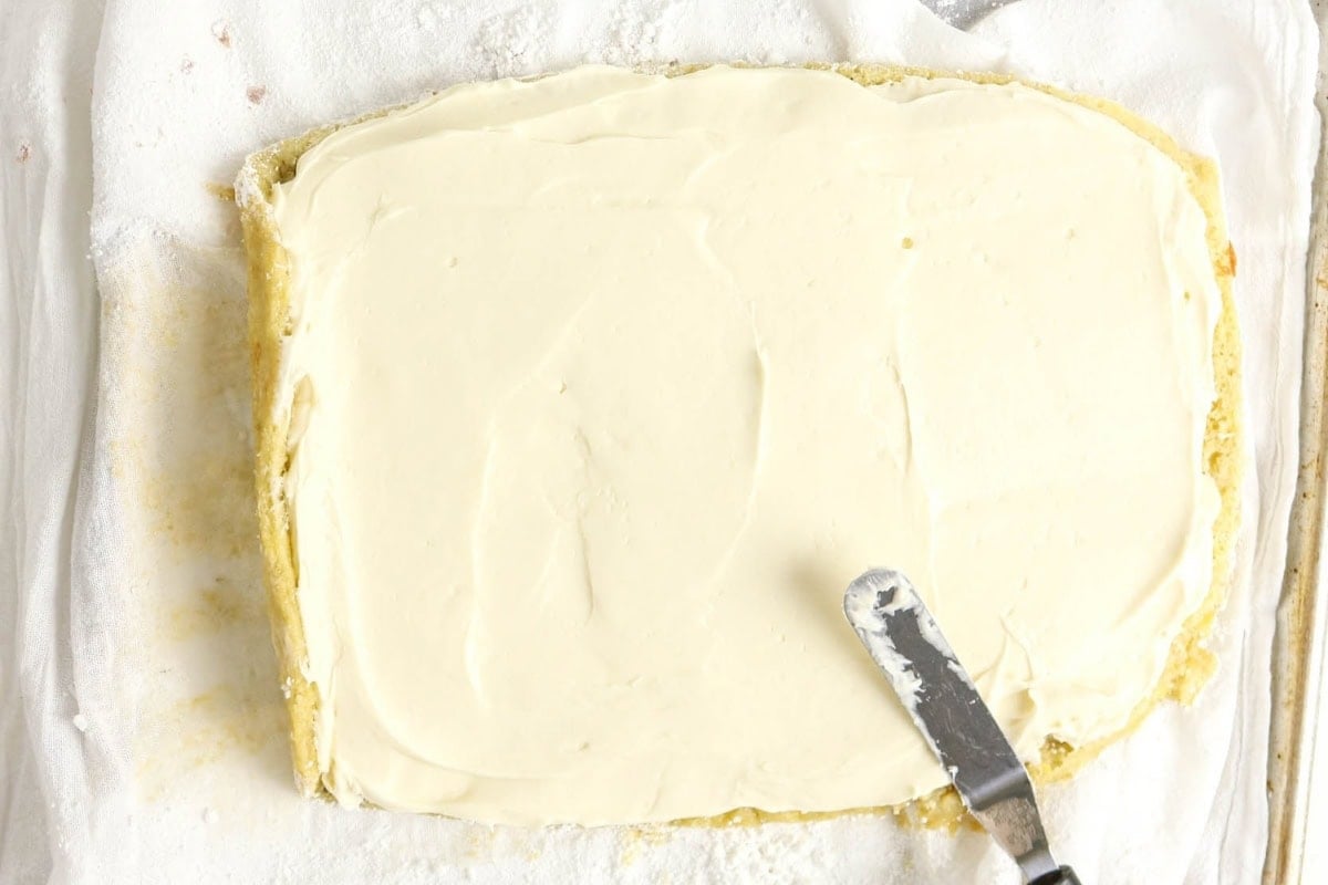 Cake being spread with white frosting using a metal spatula on parchment paper.