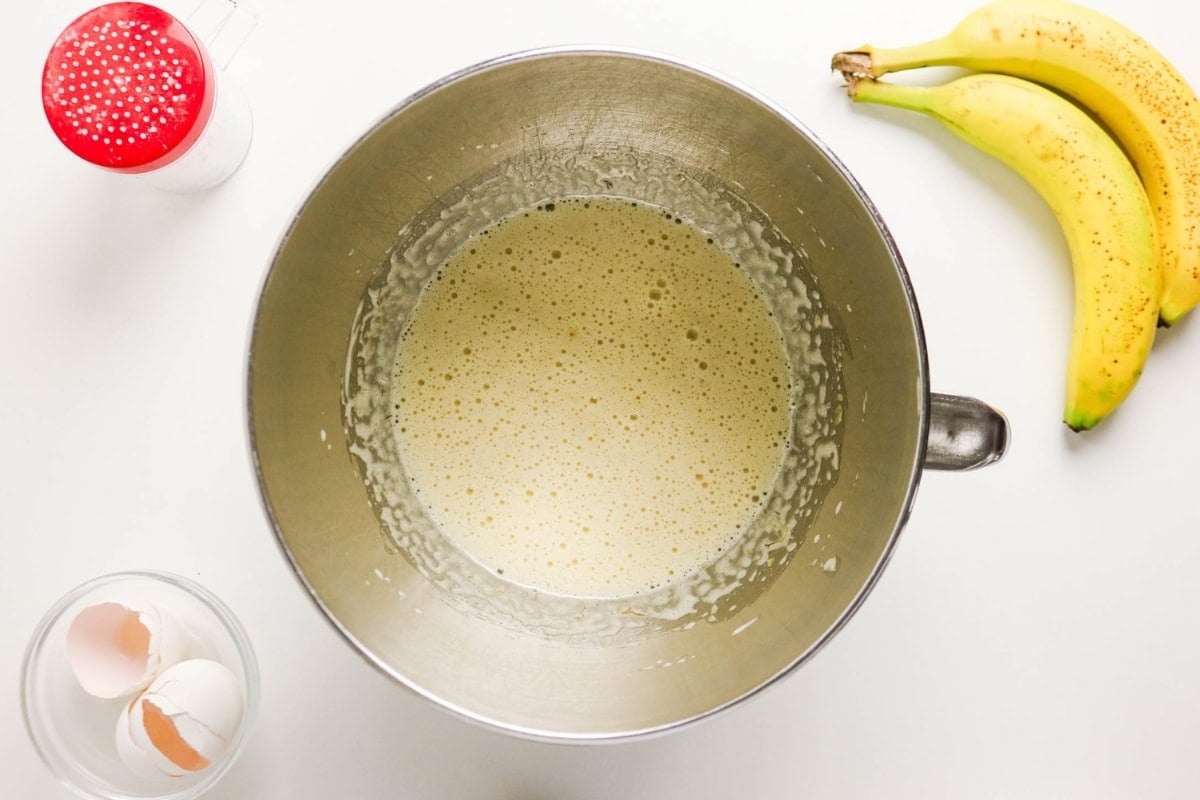 Mixing bowl of batter, bananas, eggshells, and sugar on a white surface.