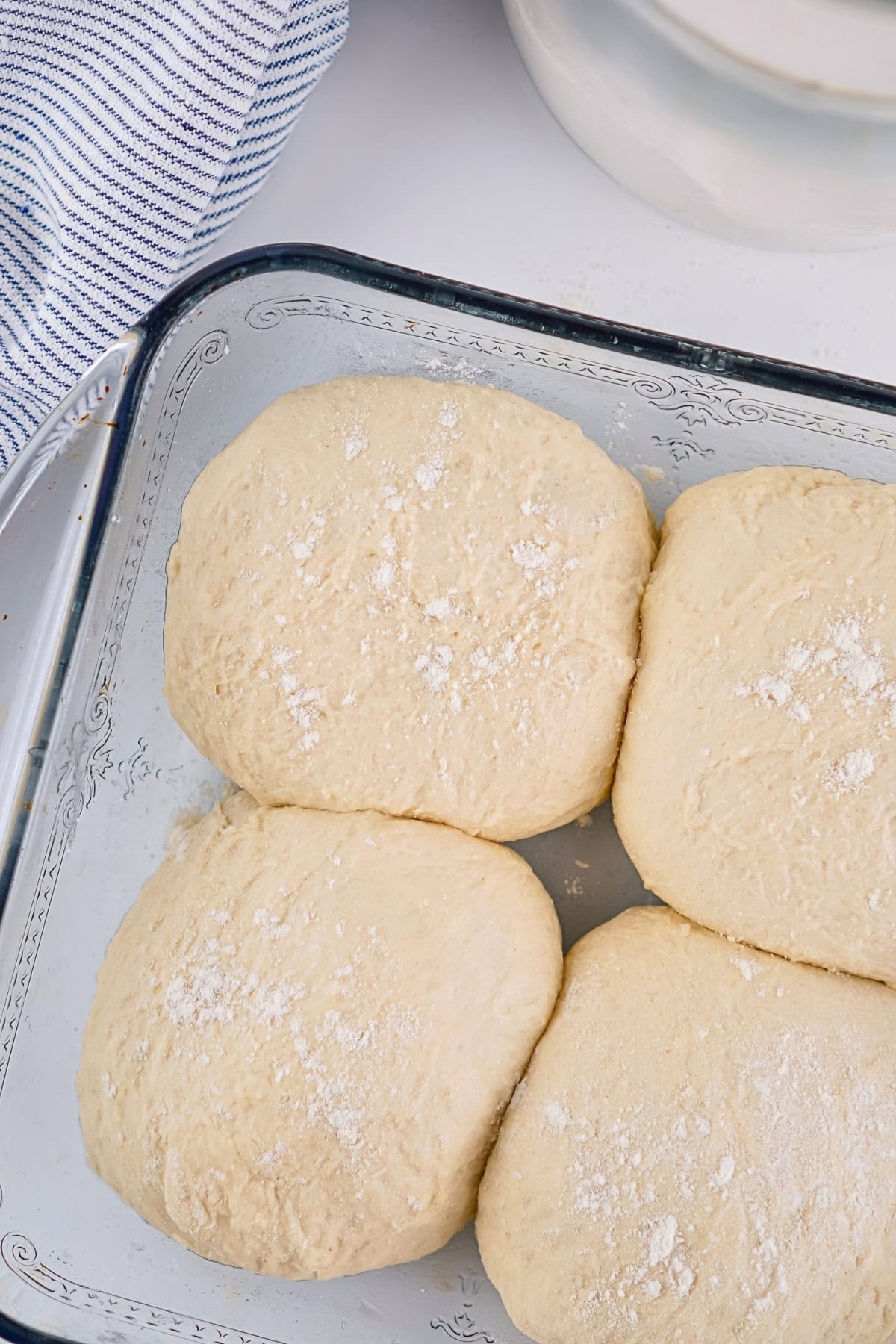 Four balls of dough with flour in a glass dish, next to a striped towel.