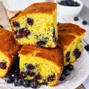 Cornbread squares with blueberries are stacked on a plate with fresh berries.