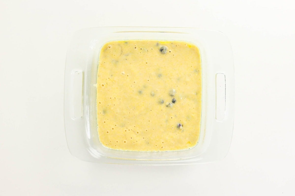 Square glass dish with yellow batter and blueberries on a white background.