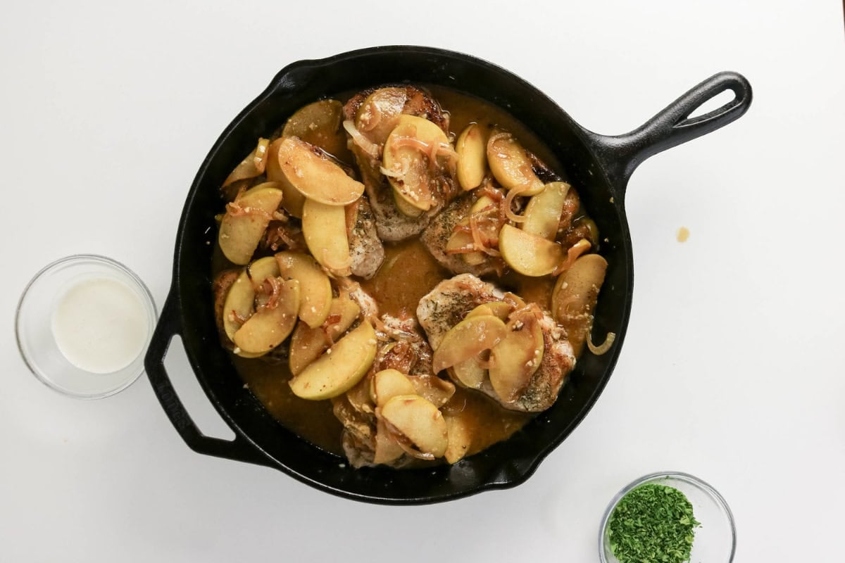 Cast iron skillet with pork chops, apple slices, and sauce, seen from above.