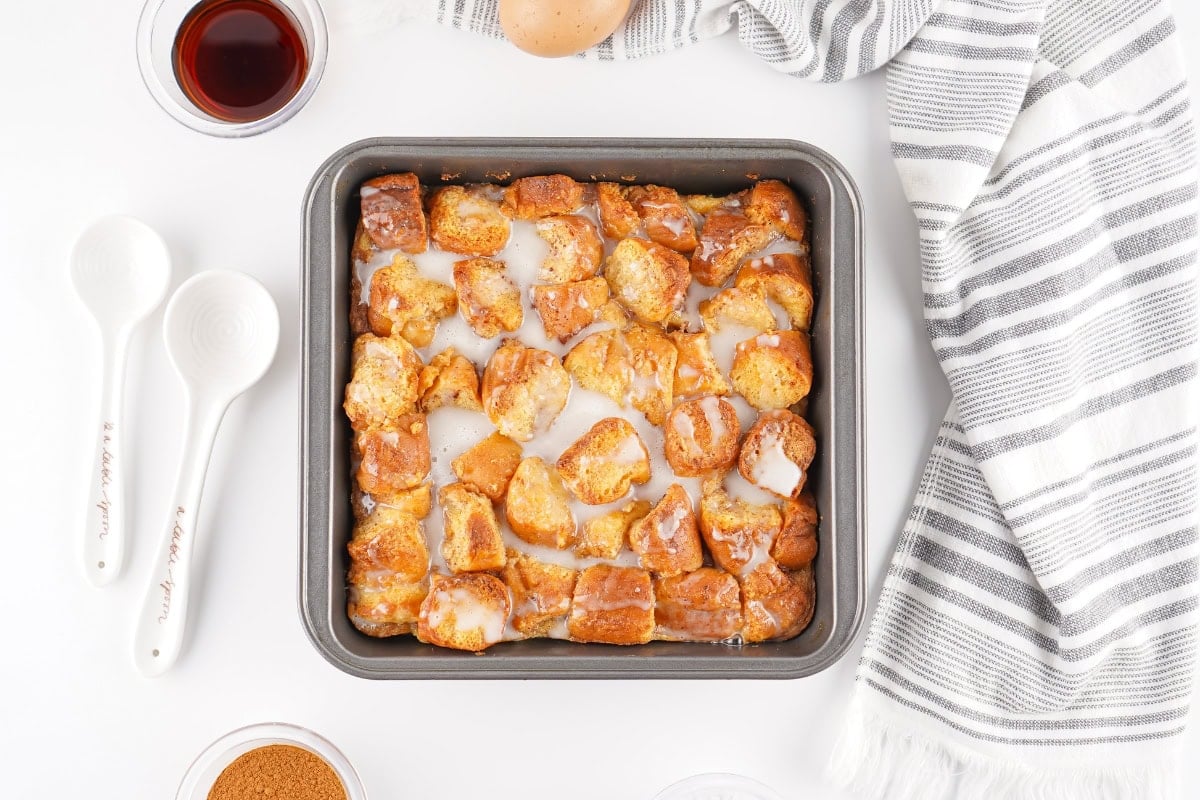 Cinnamon roll pieces with icing in a square baking pan on a white surface.