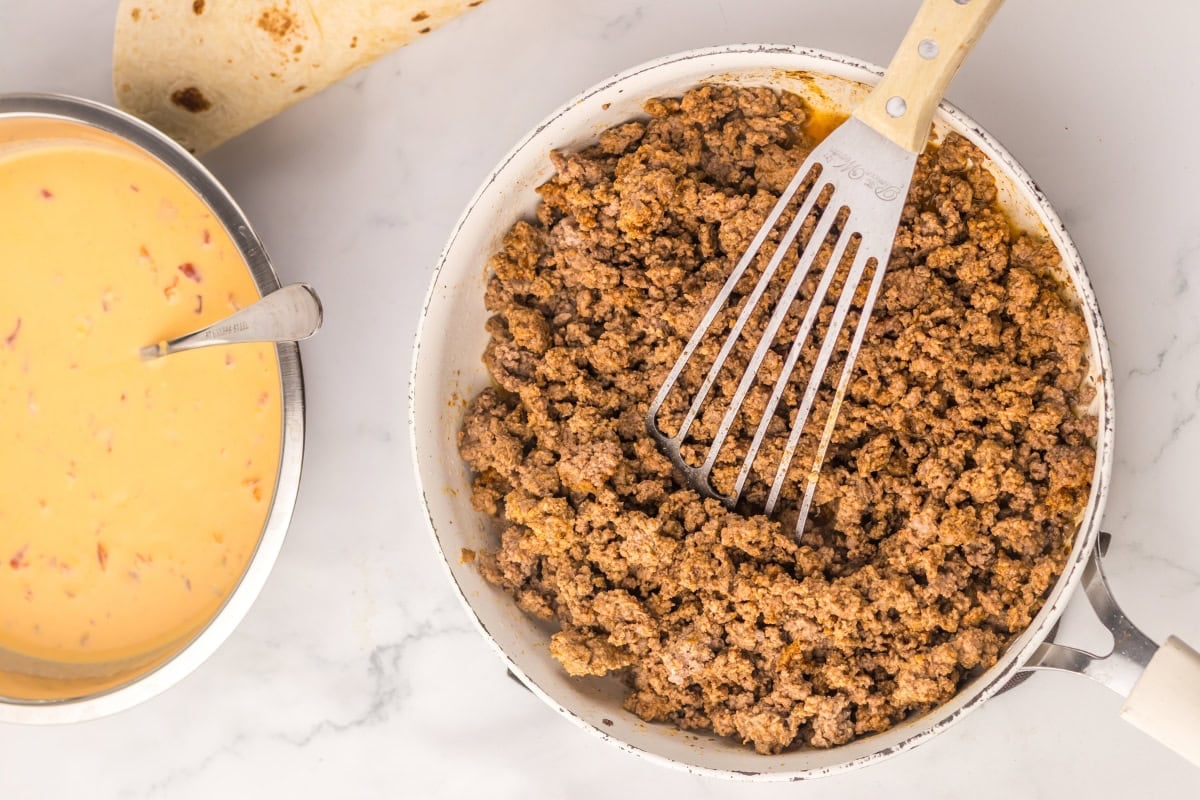 Cooked ground beef in a pan beside a bowl of cheese sauce and a tortilla.