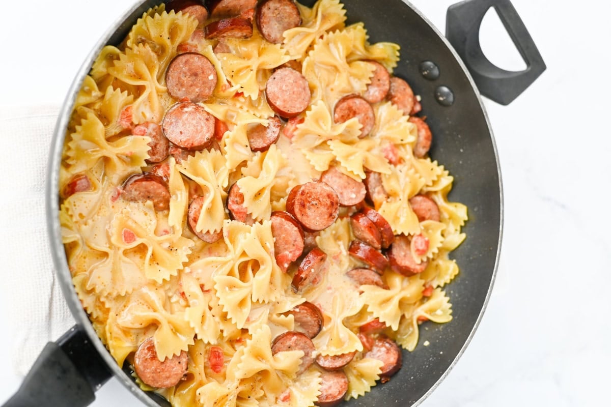 Bowtie pasta with sliced sausage in a creamy sauce in a black pan.