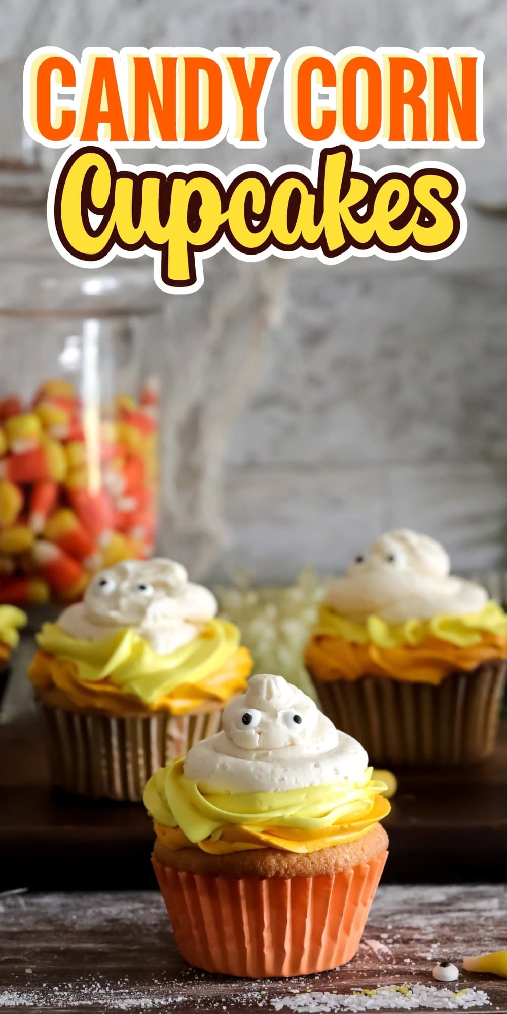 Cupcake with yellow frosting and ghost-shaped topping, candy corn in background.