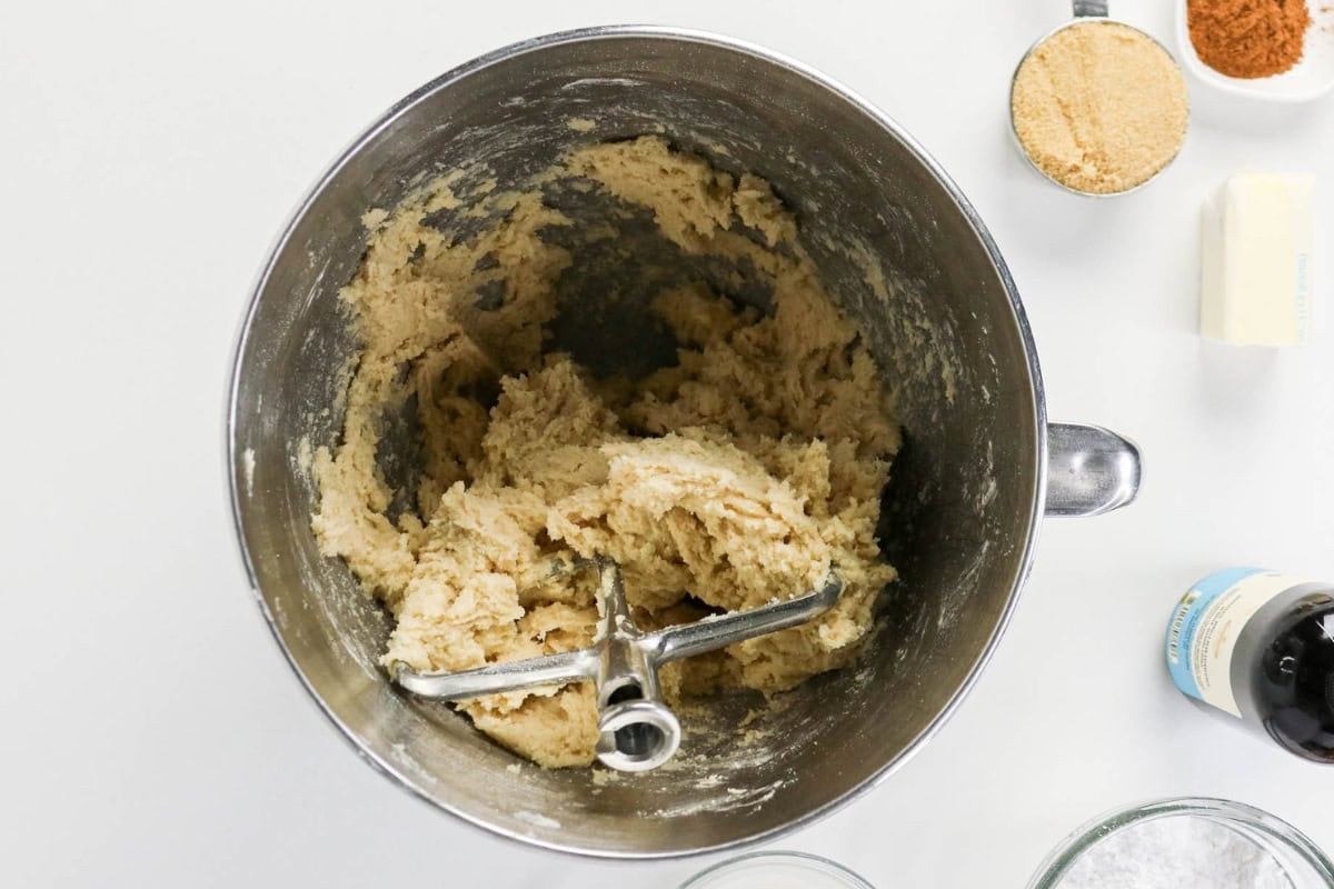 Cookie dough in a mixing bowl with ingredients around it on a white surface.