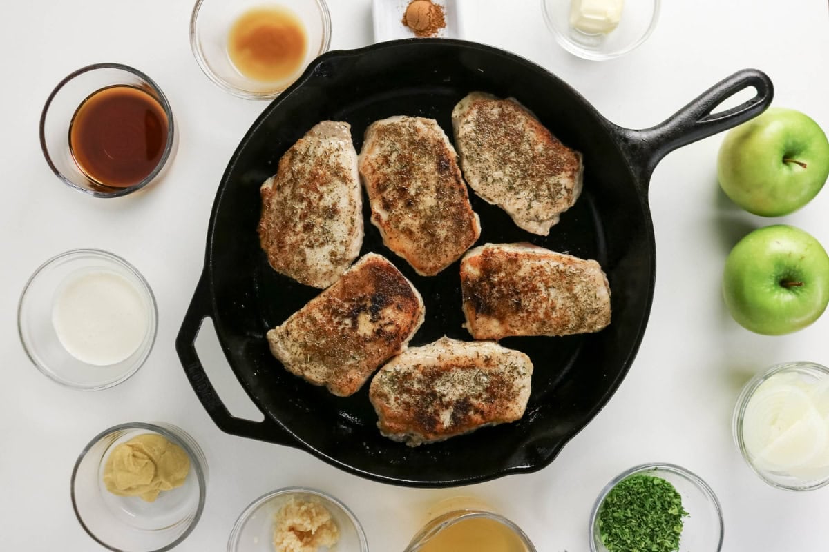 Six pork chops cook in a skillet with spices, sauces, and apples.