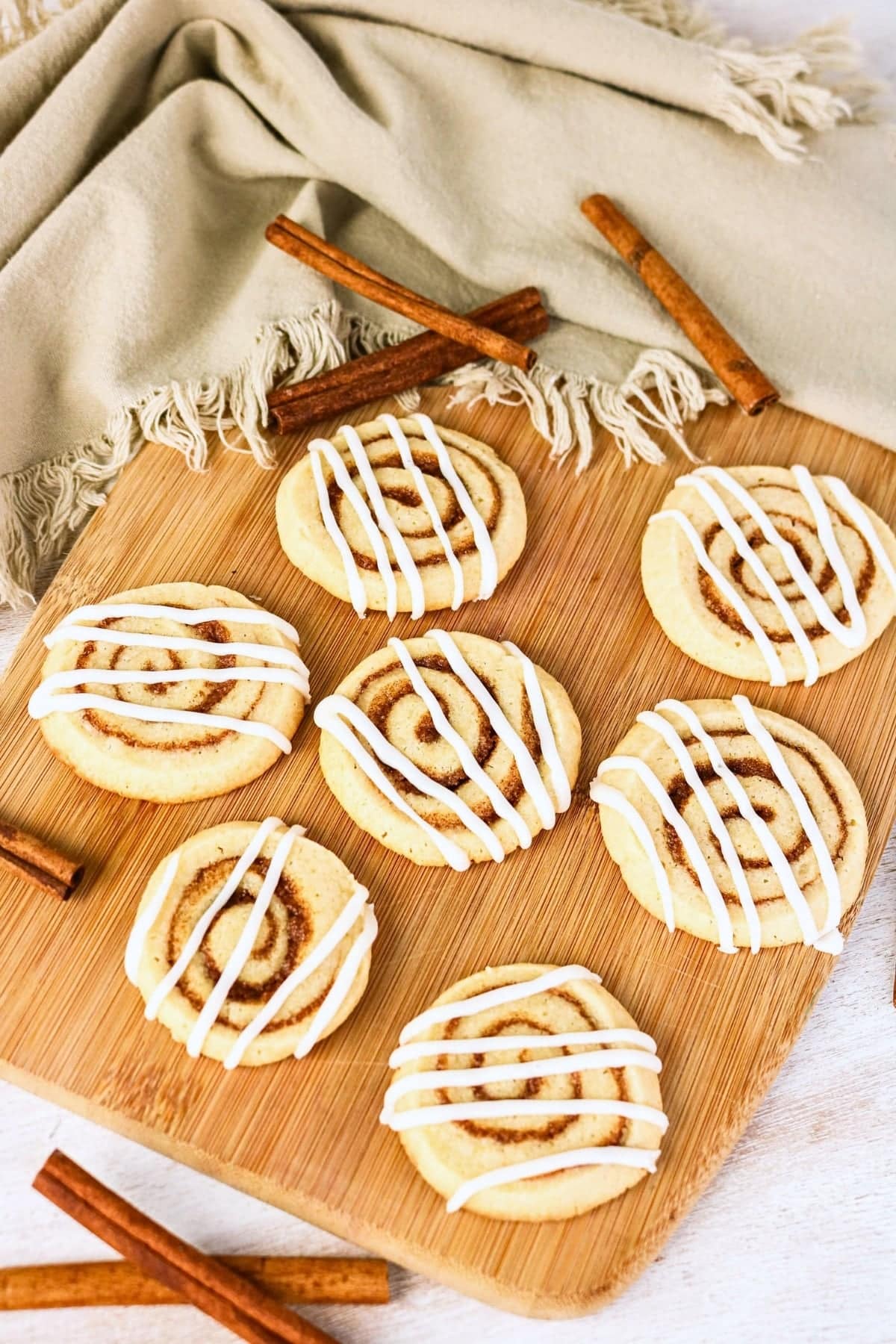 Seven cinnamon roll cookies with icing on a wooden board and cinnamon sticks.