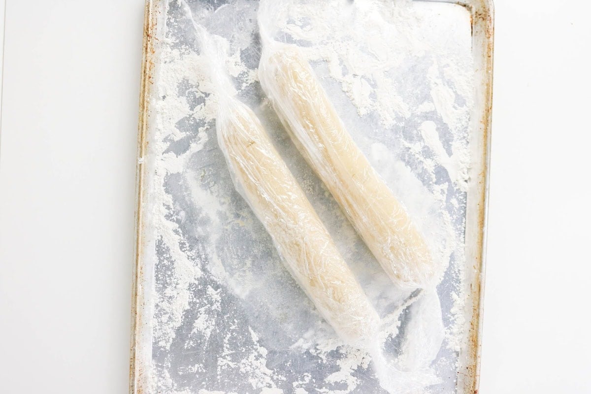 Two logs of dough wrapped in plastic wrap on a floured baking sheet.