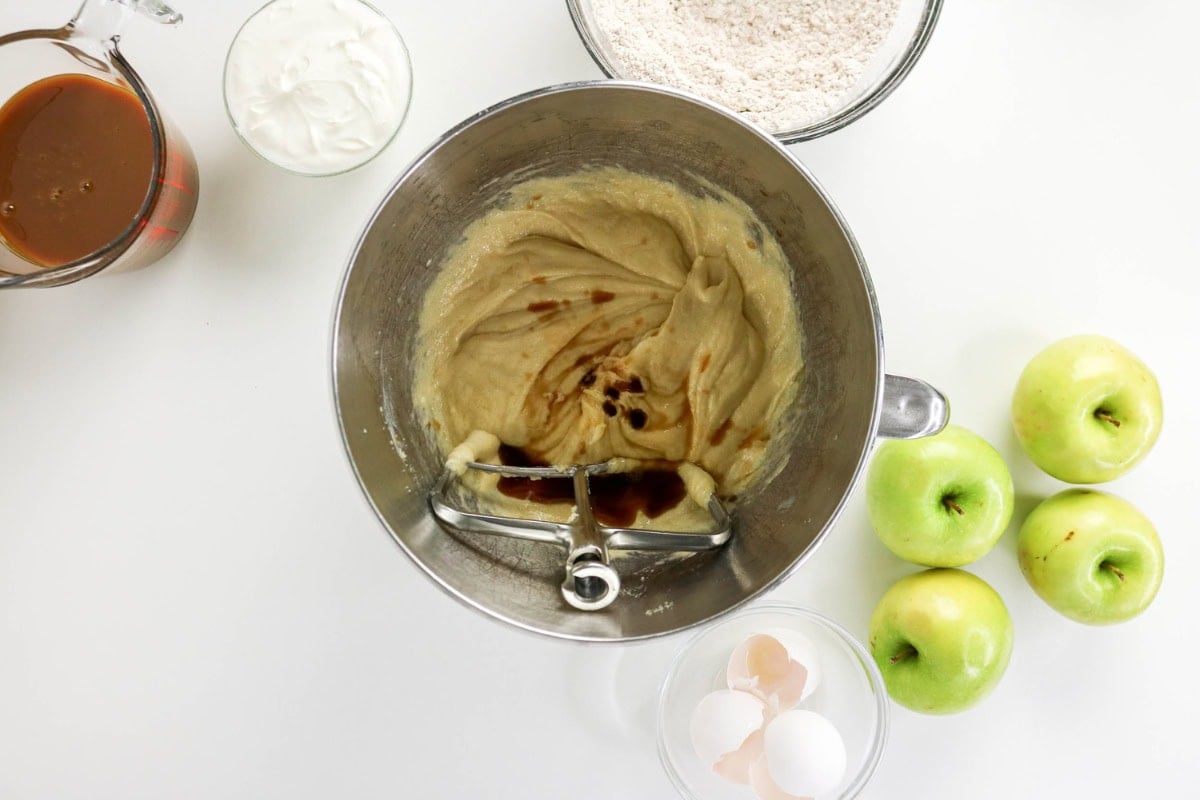 Mixer bowl with batter, vanilla, apples, eggs, flour, yogurt, and sauce nearby.