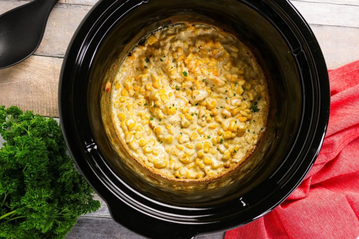 Creamed corn in a black slow cooker, with parsley and a red towel nearby.