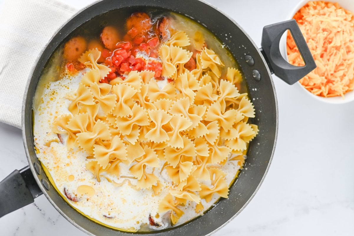Uncooked bowtie pasta, sauce, and cheese in a black pot, ready to cook.