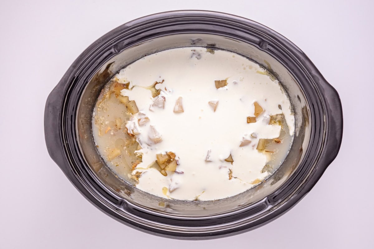 Slow cooker with cream poured over chopped potatoes and onions, ready to cook.