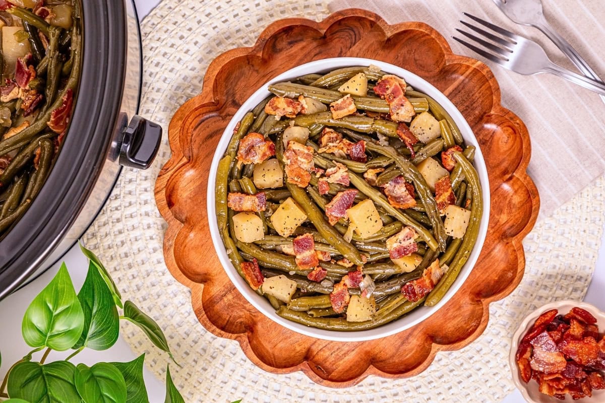 Bowl of green beans, potatoes, and bacon on a wooden plate with forks nearby.