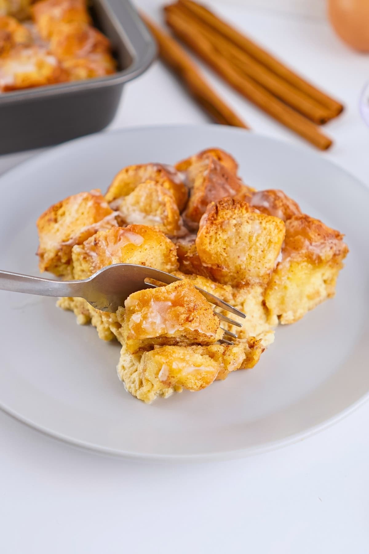 A fork lifting a piece of glazed cinnamon roll bread pudding on a gray plate.